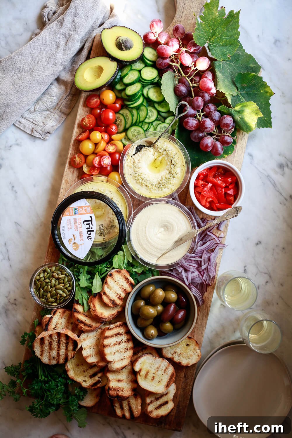 A rustic wooden board showcasing a vibrant hummus platter, featuring three containers of Tribe hummus, complemented by grilled bread, briny olives, fresh fruits, and an array of crisp vegetables, perfect for entertaining.