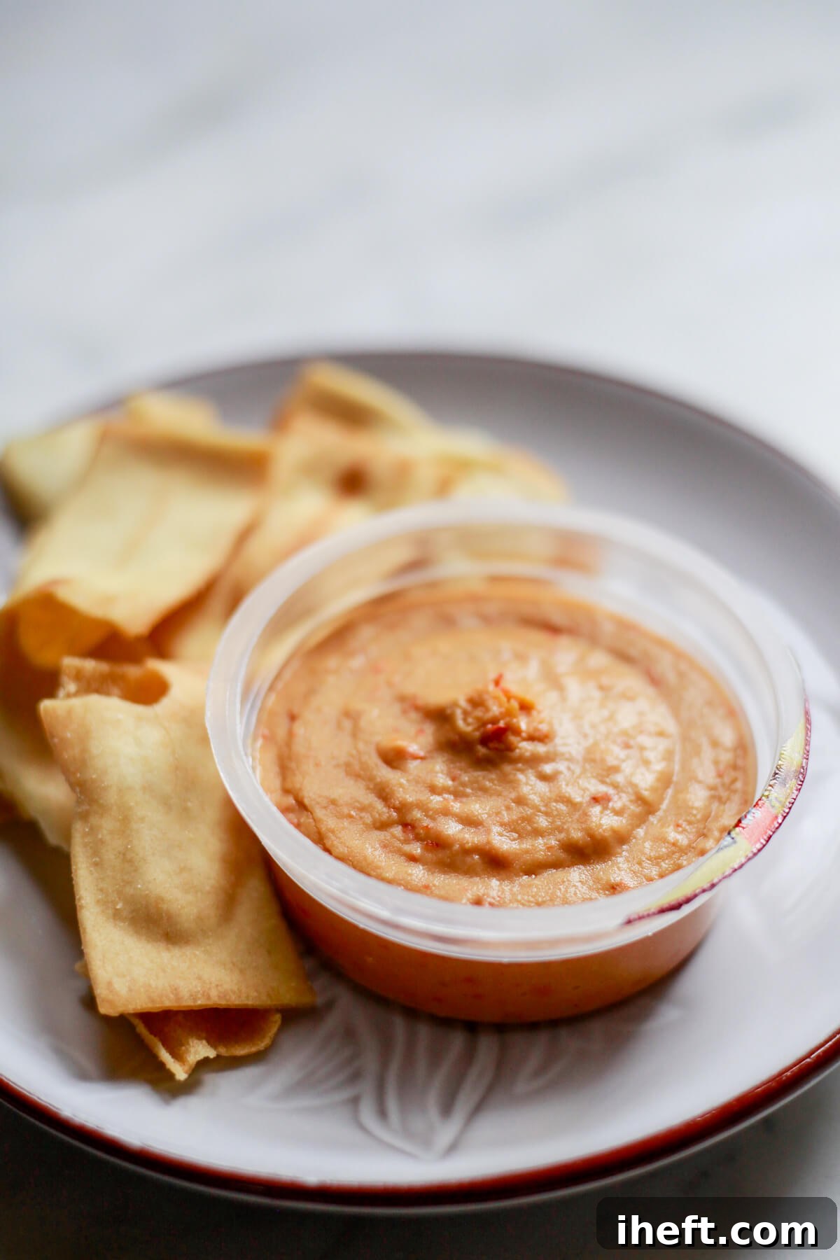 A convenient Sabra red pepper hummus snack cup, previously frozen and now thawed, presented on a white plate alongside crispy pita chips, highlighting a ready-to-eat option.