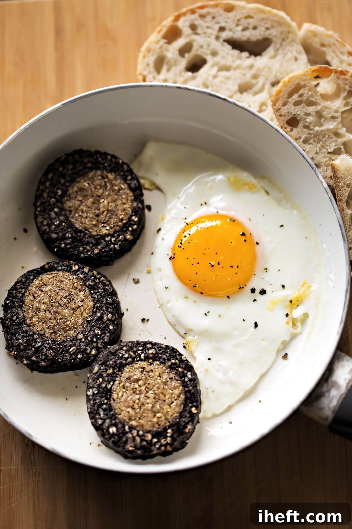 A white ceramic bowl holding a perfectly fried egg and three thick slices of both black and white pudding. This traditional Irish breakfast is elegantly served alongside a stack of sliced bread. 