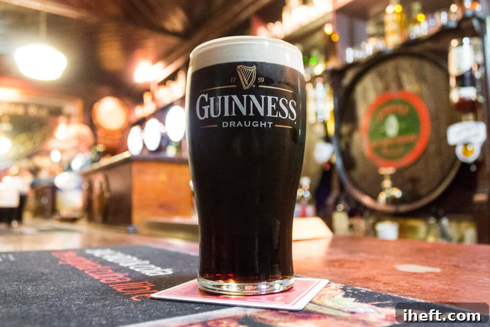 A tall, elegant glass of dark Guinness beer stands proudly on the polished counter of an Irish pub. The beer displays its characteristic rich, dark hue and a thick, creamy head of foam, invitingly presented.