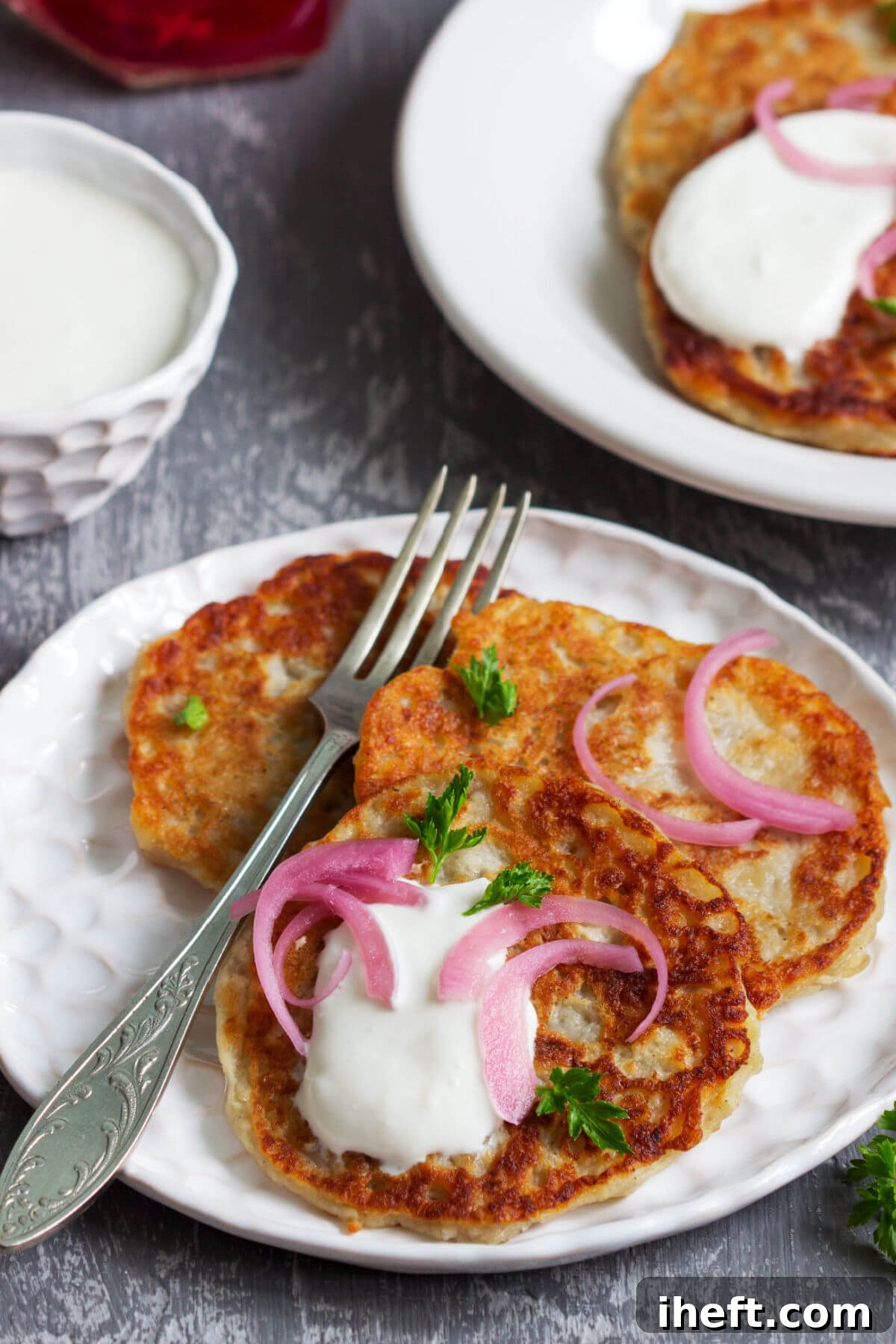 A white plate featuring three golden-brown Irish boxty potato pancakes, elegantly topped with a dollop of sour cream, finely chopped pickled onions, and fresh parsley. A vegetarian friendly Irish food. 