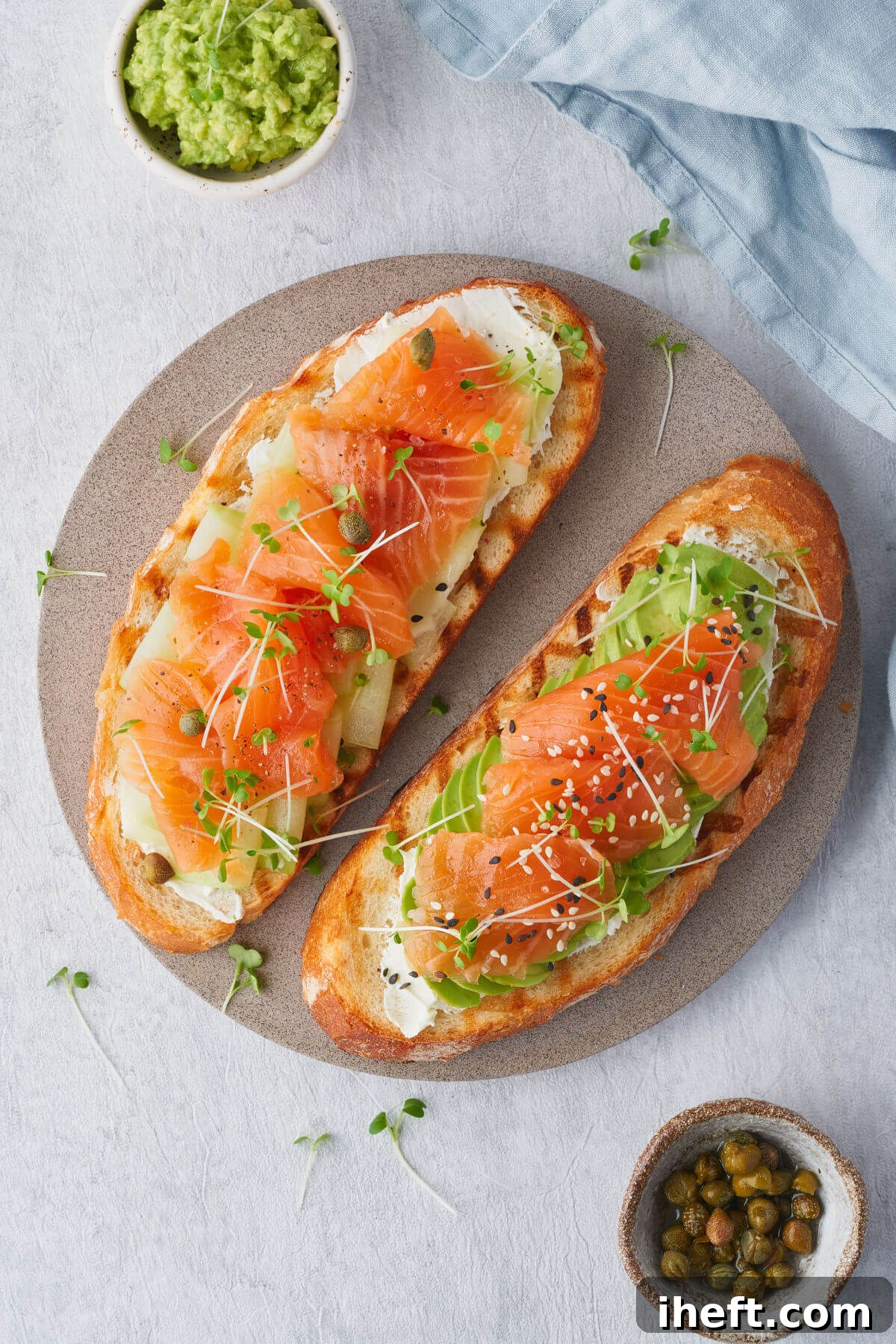 Two slices of perfectly toasted bread generously topped with creamy cream cheese, fresh avocado slices, and delicate smoked salmon, garnished with vibrant microgreens. A small bowl of briny capers sits elegantly to the side on a clean white backdrop. 