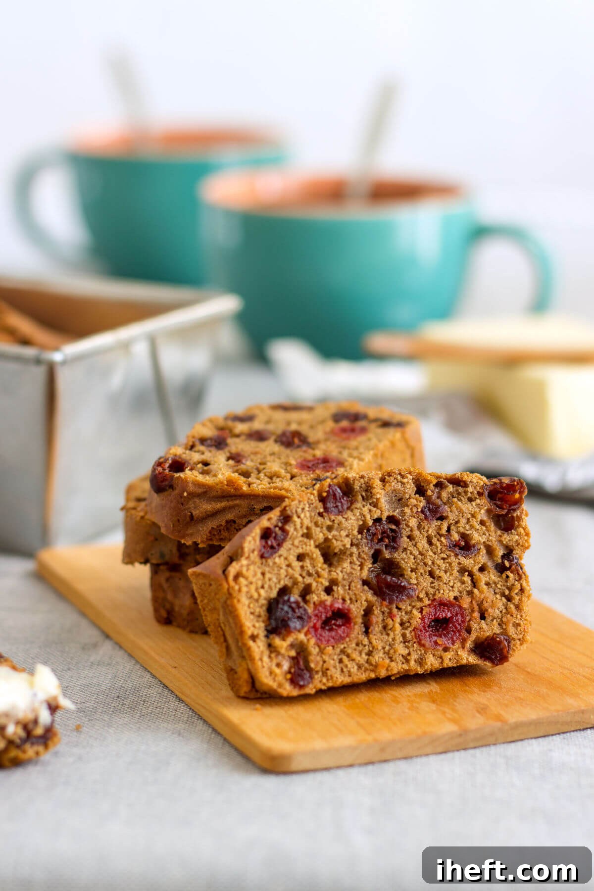 A freshly baked loaf of homemade Irish barmbrack bread is presented on a wooden cutting board, with a loaf pan and coffee mugs subtly blurred in the background. This rustic brown quickbread reveals slices studded with bright red cherries, highlighting its fruity appeal. 