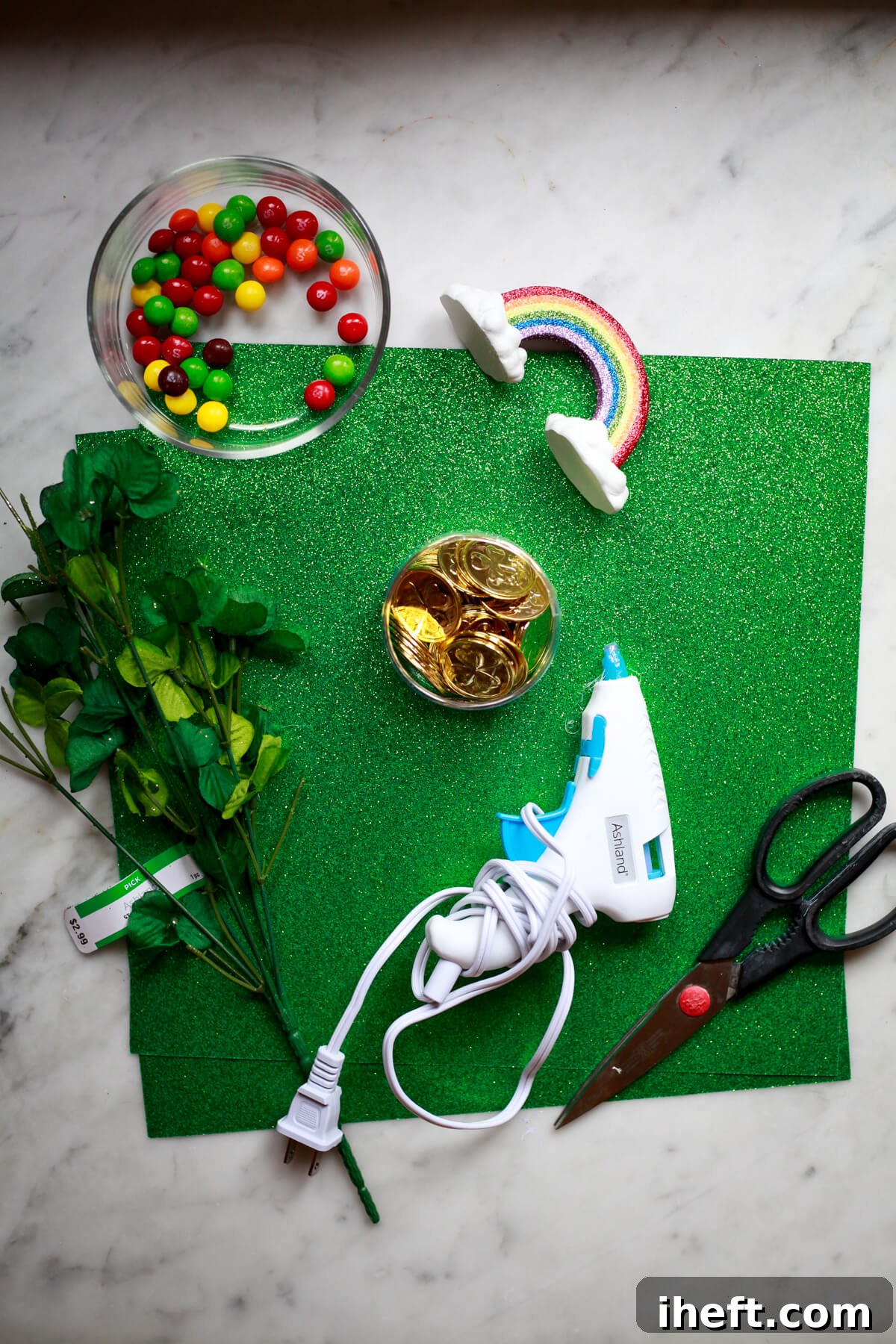 Green paper, skittles, a rainbow, gold coins, faux clovers, a glue gun, and scissors on a marble countertop to show leprechaun trap supplies. 