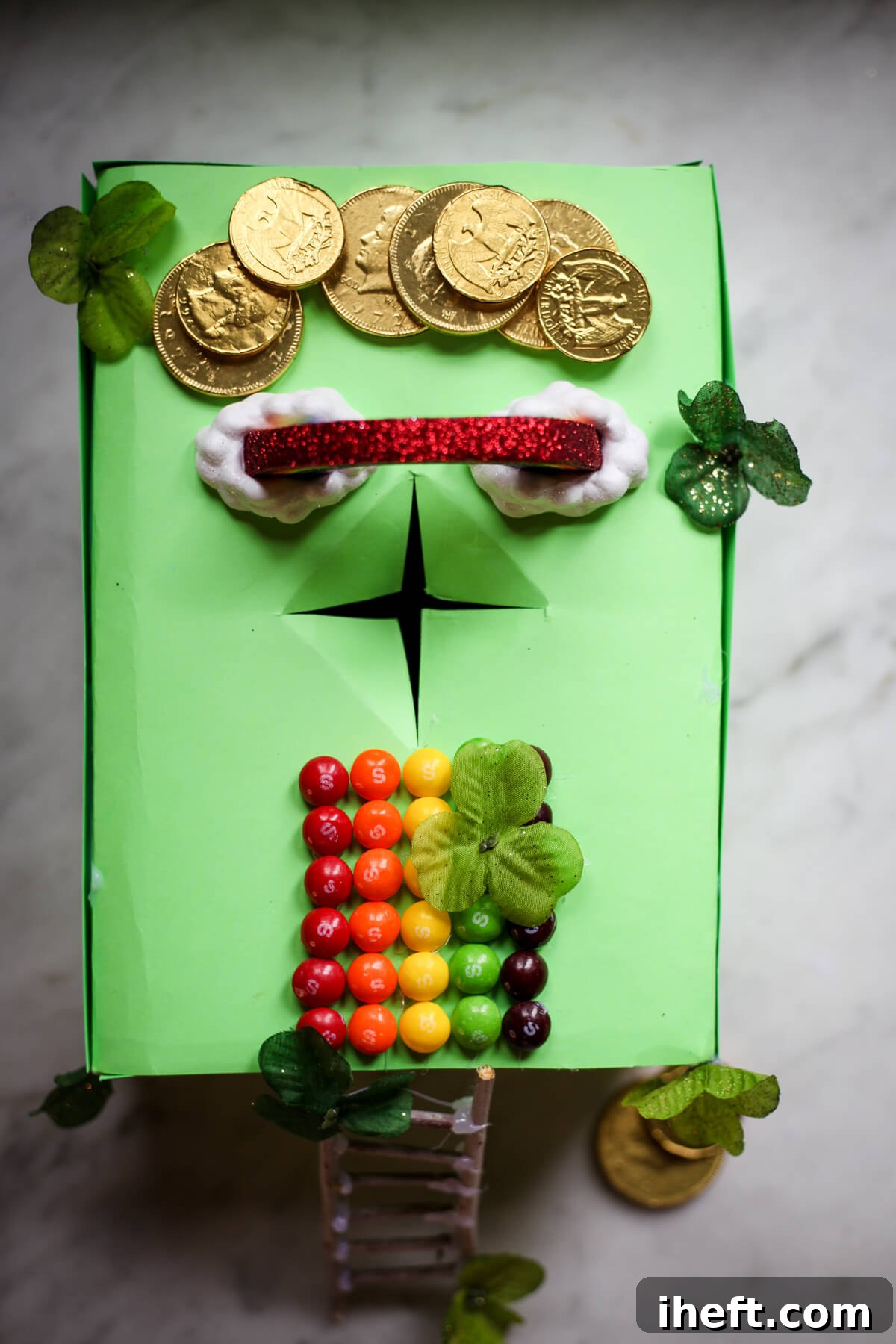 An easy leprechaun trap idea made with a shoebox covered with green paper. There is a path made with skittles, gold coins, a rainbow, and stick ladder. 