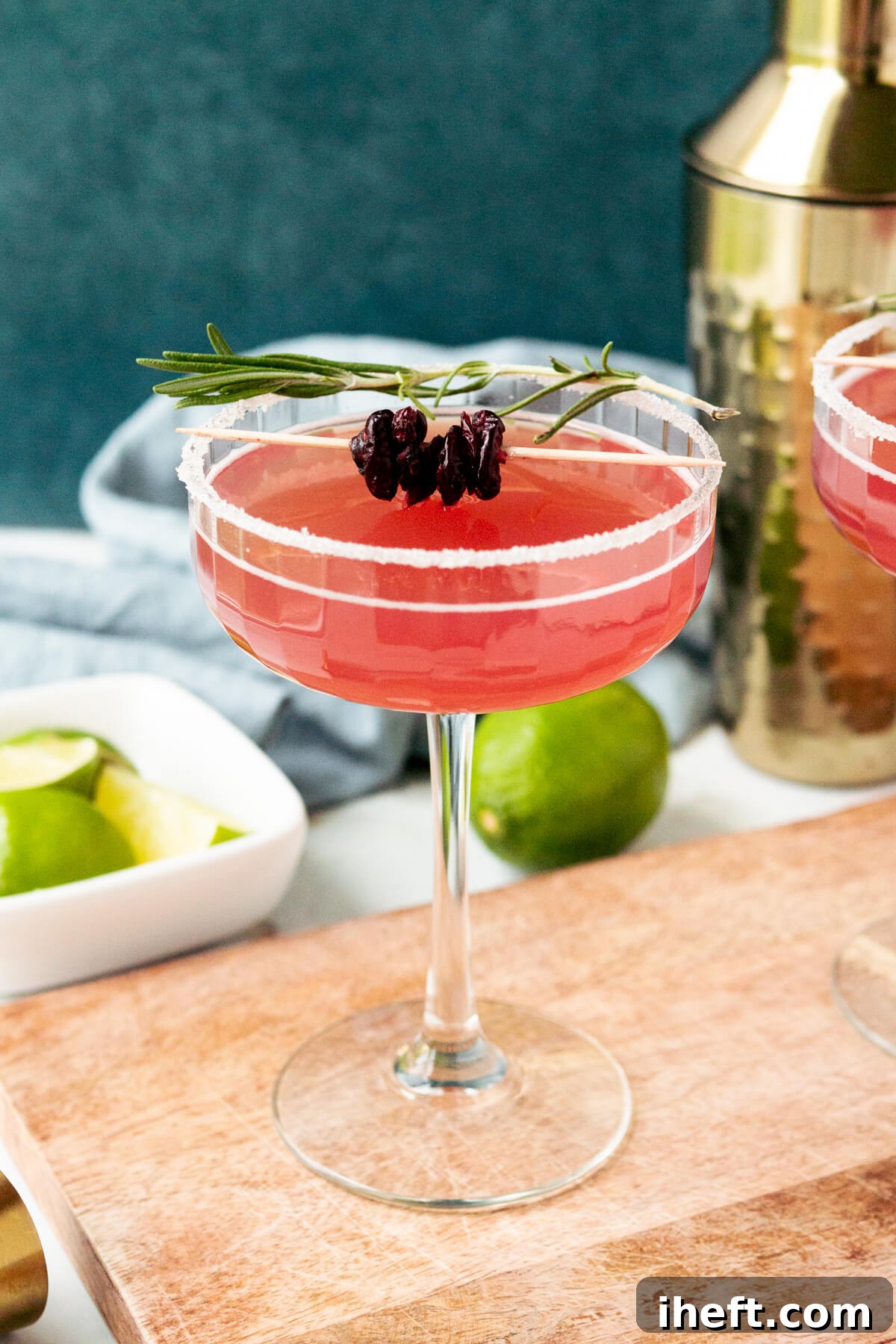 A pink Cadillac Margarita in a coupe glass garnished with dried cranberries and a sprig of rosemary. 