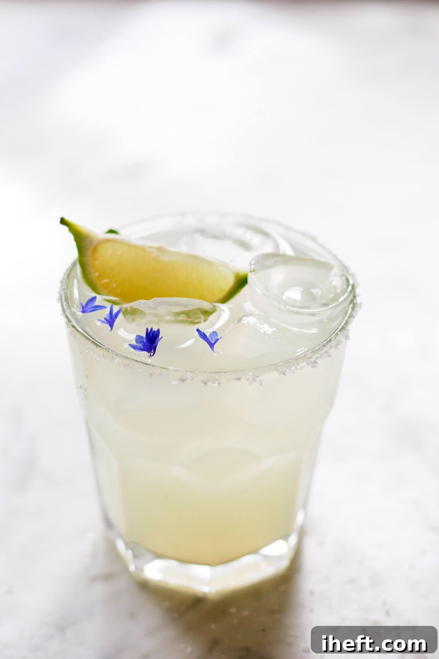 A Cadillac Margarita on the rocks garnished with a lime wedge and bachelor buttons flowers. 