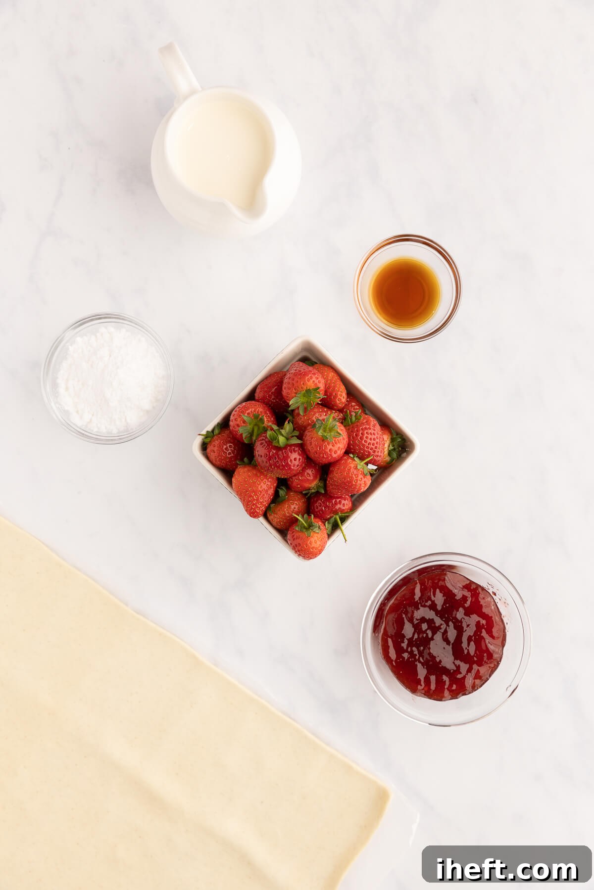 The ingredients for making a Napoleon dessert sit on a white marble countertop. These include heavy cream, vanilla, strawberries, strawberry jam, puff pastry, and powdered sugar. 