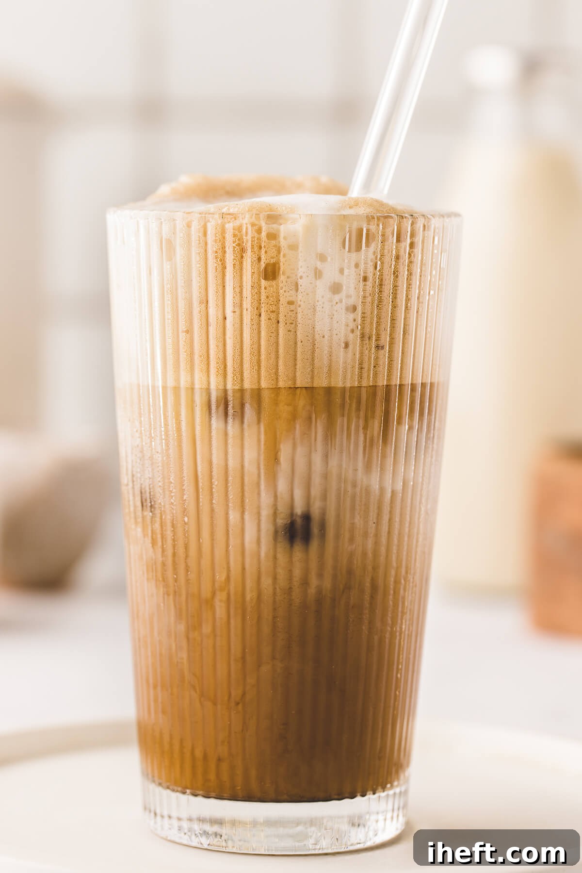 A close-up photo of a Brown Sugar Oatmilk Shaken Espresso in a tall glass with ice, frothy cold foam, and a glass straw. This homemade iced shaken espresso recipe sits on a white kitchen counter. 