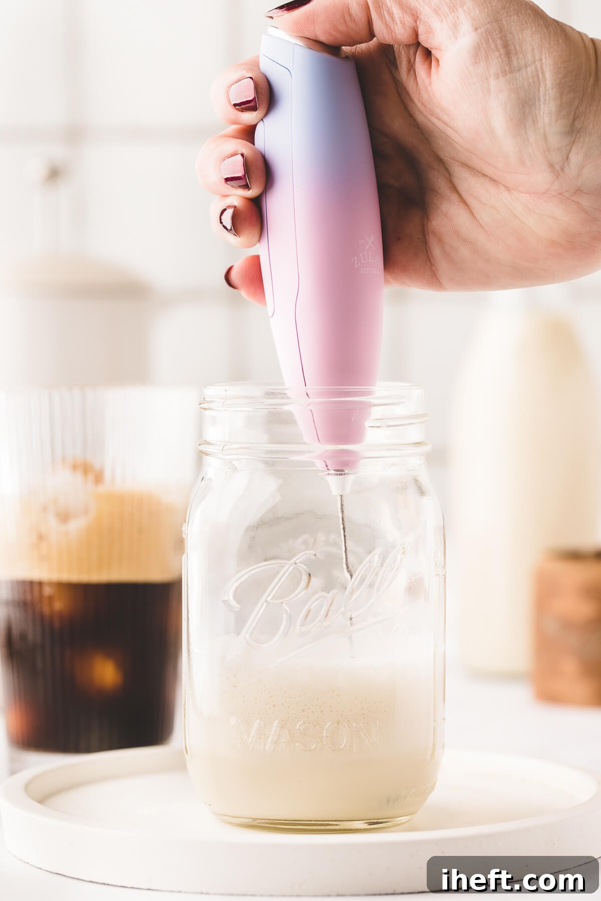A pink milk frother makes cold foam out of oat milk in a mason jar. Shaken espresso in a glass can be seen in the background.