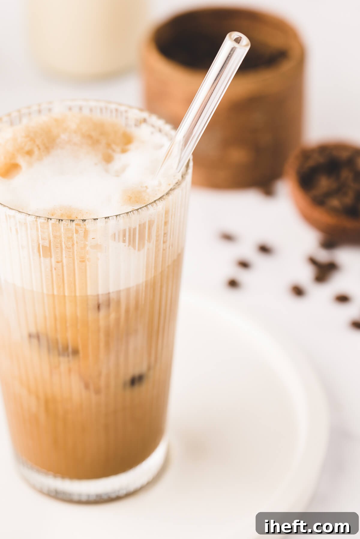 An oatmilk iced shaken espresso with cold foam in a tall glass with glass straw. Coffee beans in the background on a white counter. 