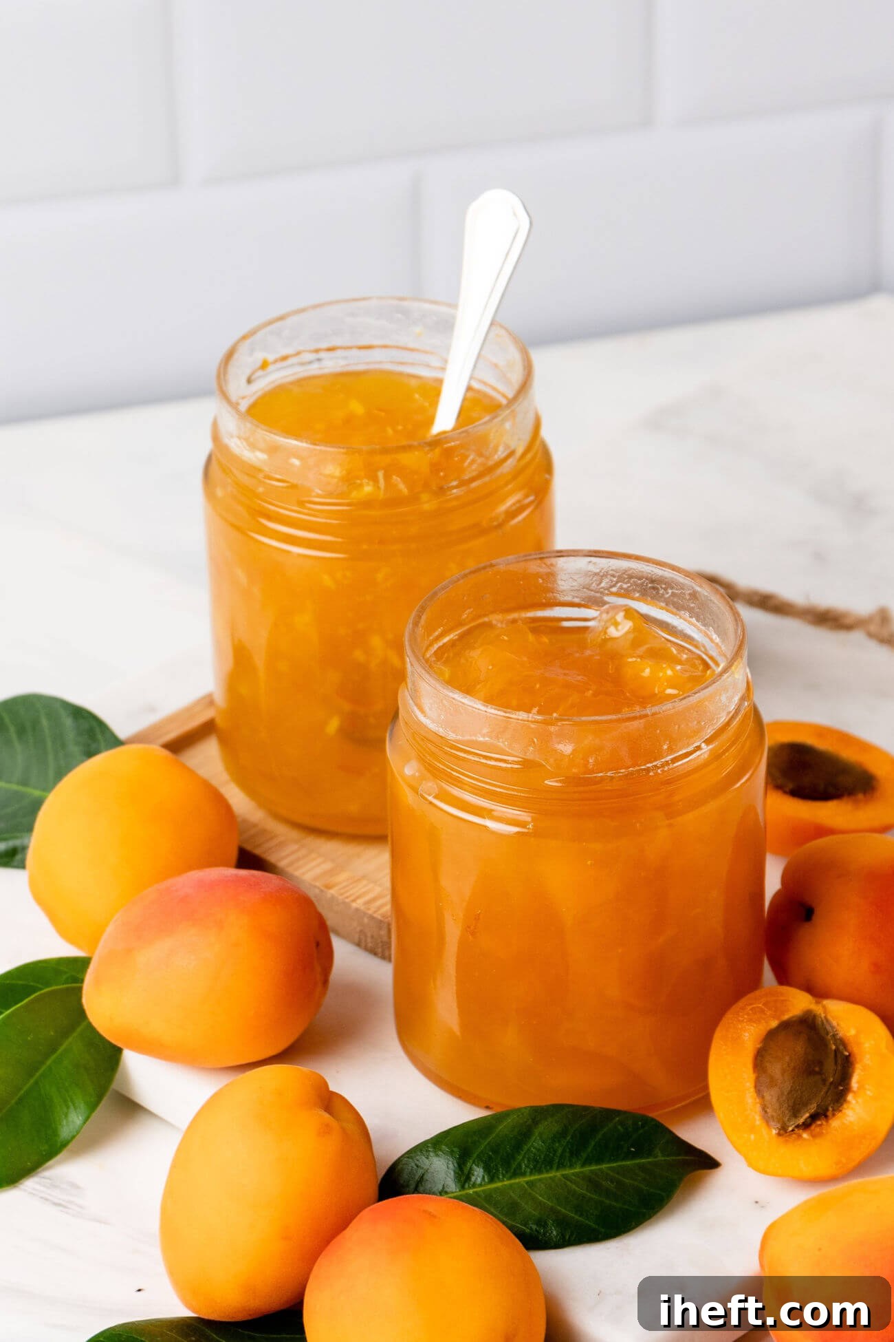 Two glass jars filled with homemade apricot jam on a white counter. Fresh apricots and leaves also sit on the counter. 