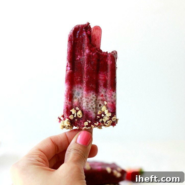 Hand holding a purple acai, chia, and granola popsicle with a bite taken out of it, showing the layers and fruit pieces. 