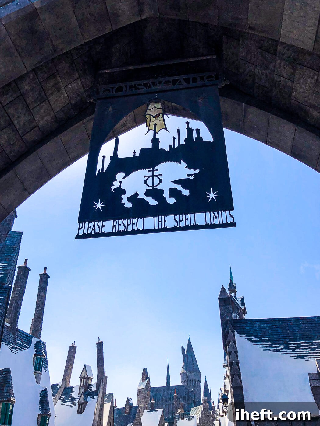 The grand stone entrance to The Wizarding World of Harry Potter™ at Universal Studios, adorned with a Hogwarts sign that politely reminds visitors to "please respect the spell limits." Snow-capped buildings loom majestically in the background.