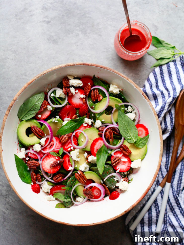 A vibrant Strawberry Spinach Salad with Strawberry Vinaigrette. The image showcases fresh baby spinach, sliced strawberries, crumbled feta, red onion, avocado, basil, and candied pecans, all dressed in a homemade strawberry balsamic vinaigrette.