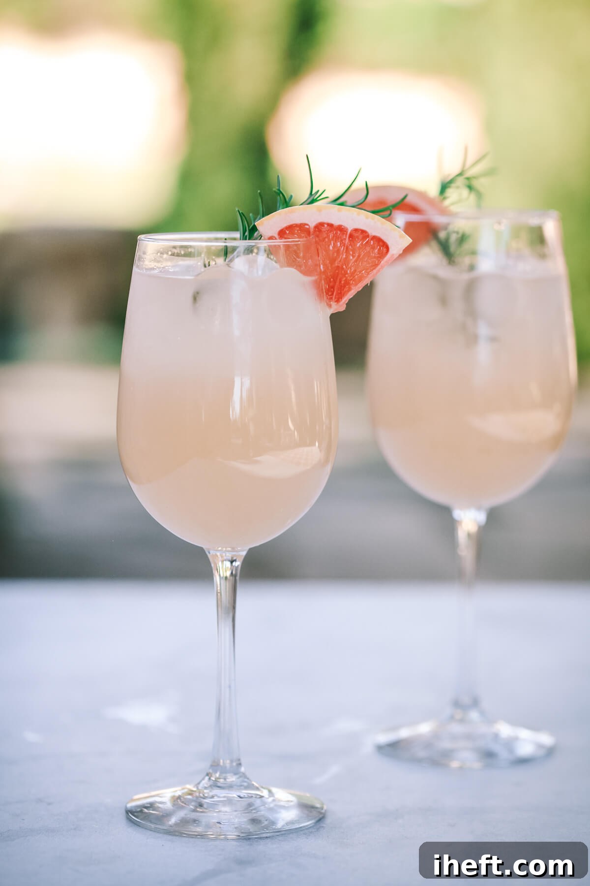 Two elegant wine glasses filled with a pale pink grapefruit beverage, garnished with fresh pink grapefruit wedges and sprigs of fragrant rosemary. The drinks suggest a sophisticated and refreshing option for a pink-themed party.