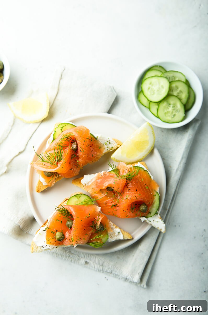 Three pieces of toasted bread adorned with creamy white cheese, crisp cucumber slices, briny capers, and fresh dill, presented on a pristine white plate. This dish serves as an excellent visual example of Smoked Salmon Cucumber Toast.