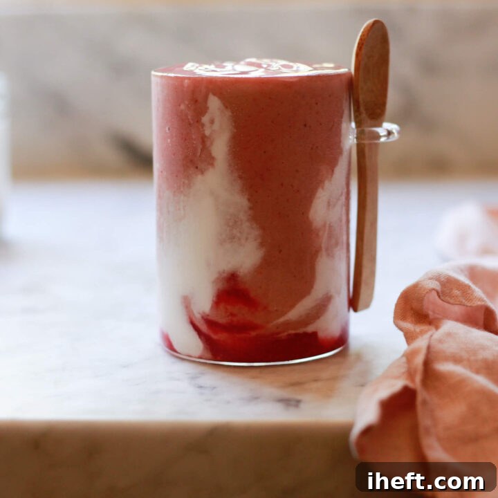 A pink strawberry Hailey Bieber smoothie copycat with coconut creme swirls sits on a marble kitchen countertop.