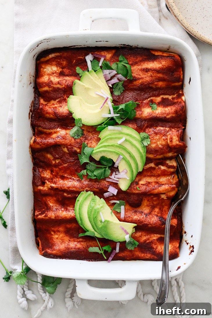 A vibrant pan of red enchiladas, beautifully garnished with fresh sliced avocado, chopped cilantro, and thinly sliced red onion. This image highlights the appealing and fresh presentation of these delicious black bean and corn enchiladas, emphasizing their vegetarian and vegan-friendly nature.