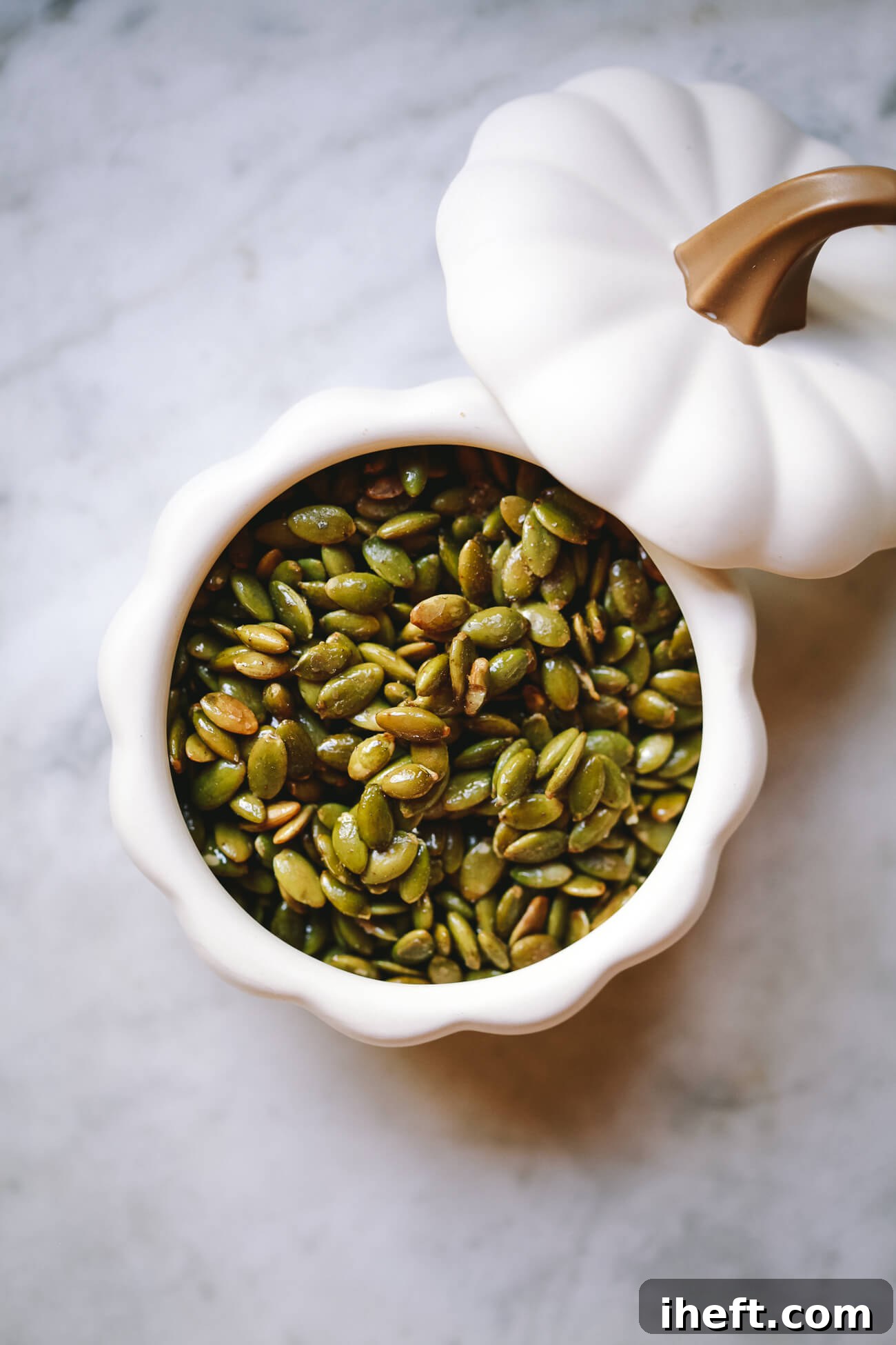 A white pumpkin-shaped ceramic bowl filled with roasted pepitas (pumpkin seeds).