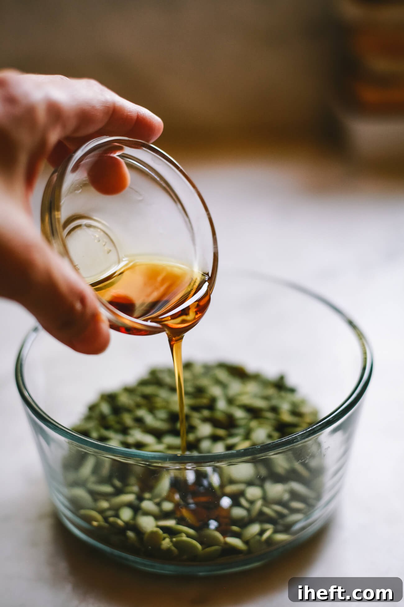 A small bowl of maple syrup is poured over raw pepitas in a glass bowl. This is the process for making a maple glazed roasted pepitas recipe.