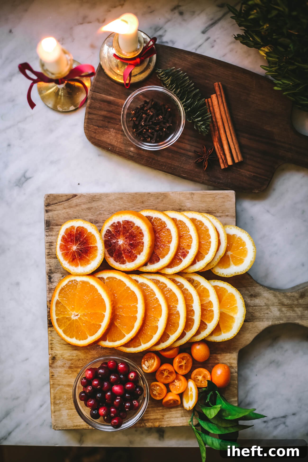 Sliced oranges and kumquats on a wooden cutting board with fresh cranberries, cloves, star anise, cinnamon sticks, and pine sprig. Lit candles in gold candle stick holders in the corner. 