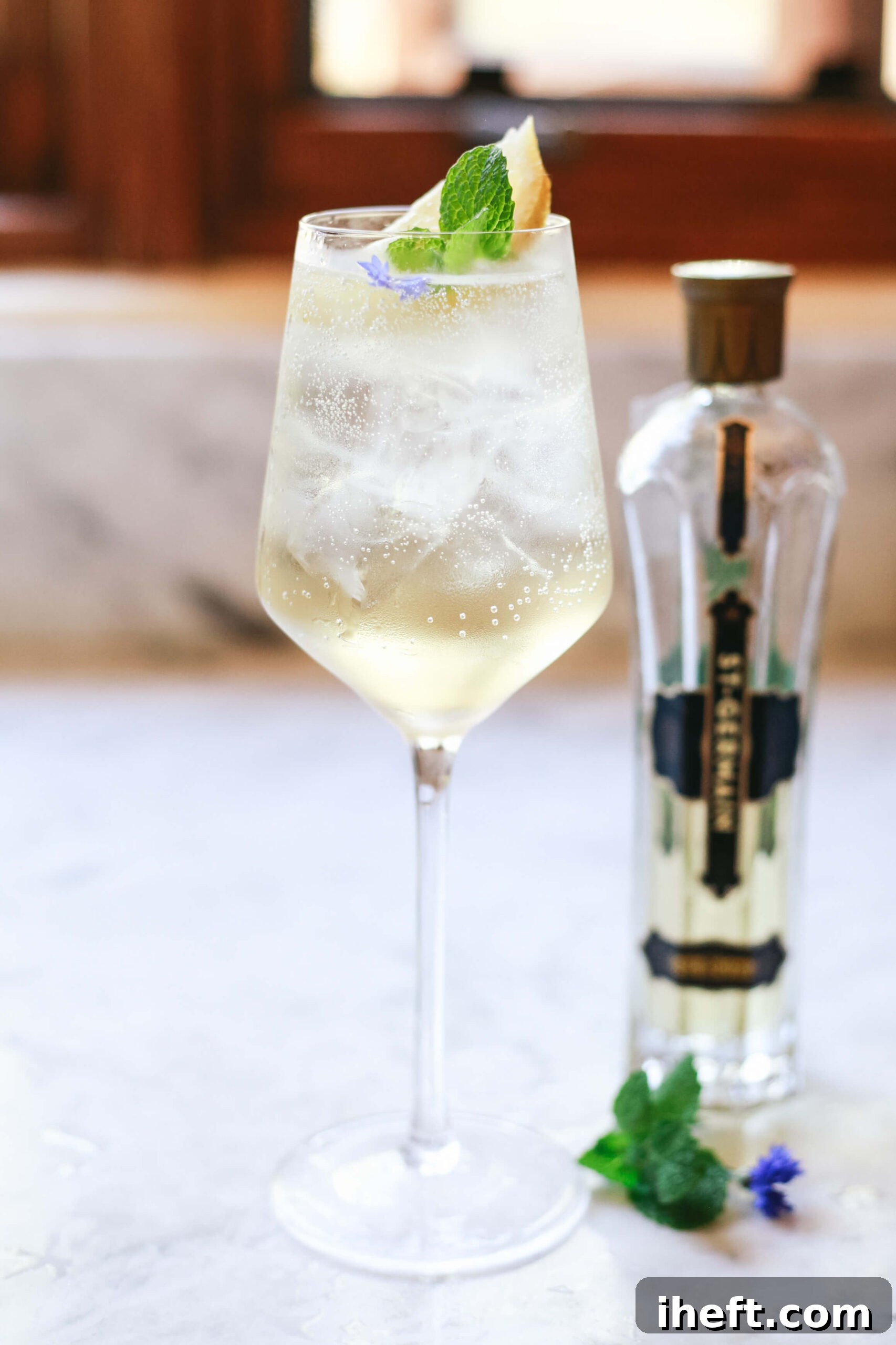 Glass of Hugo Spritz cocktail with ice, mint, and lime garnish, served in a wine glass with a bottle of St-Germain elderflower liqueur in the background.