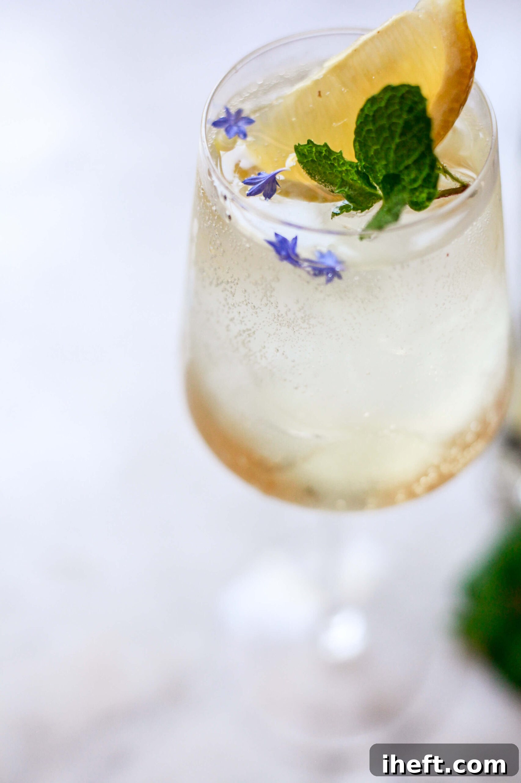Close-up of a Hugo Spritz cocktail with lemon wedge, fresh mint, and edible purple flowers floating on ice in a wine glass.