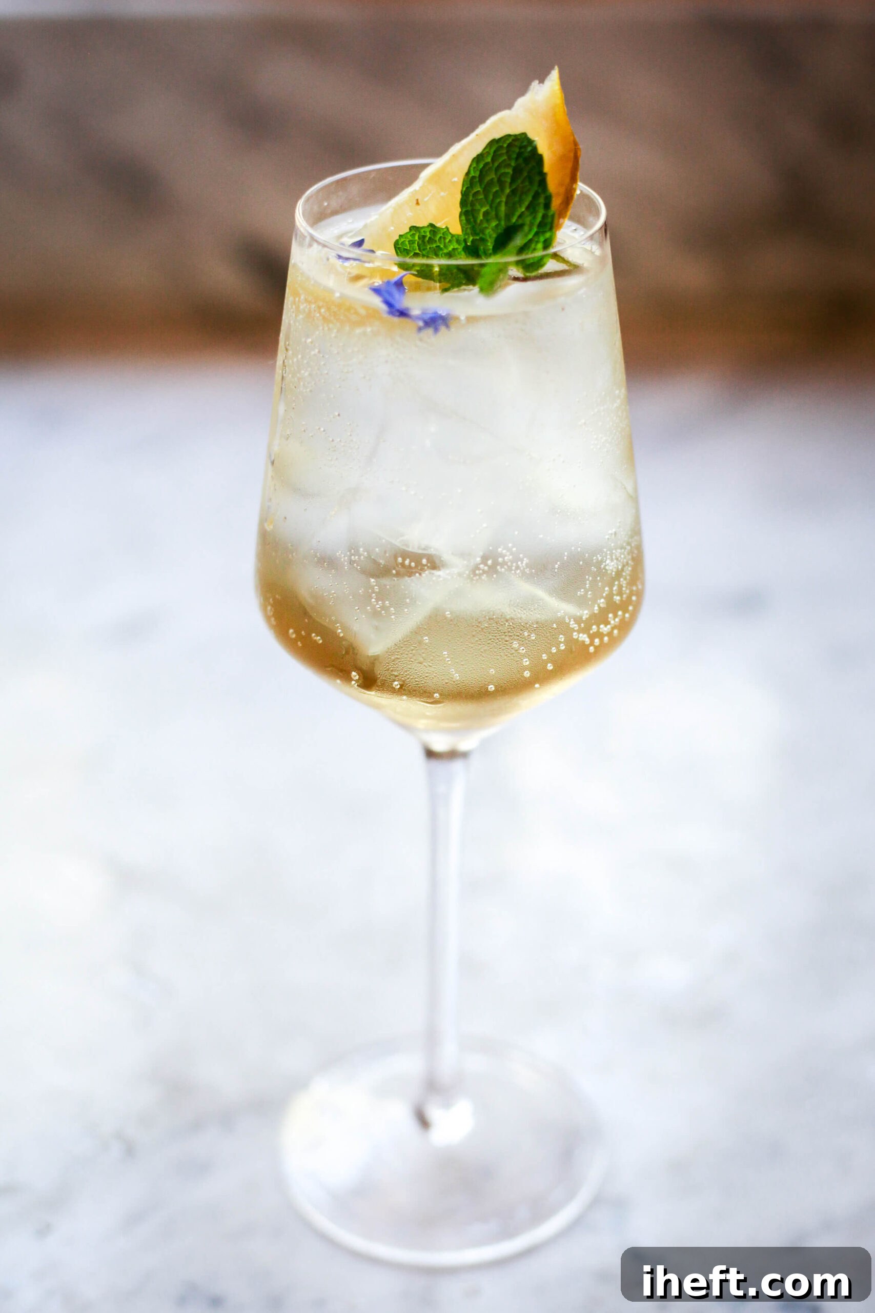 Tall wine glass filled with Hugo Spritz cocktail, garnished with lemon wedge, mint sprig, and purple flowers on ice.