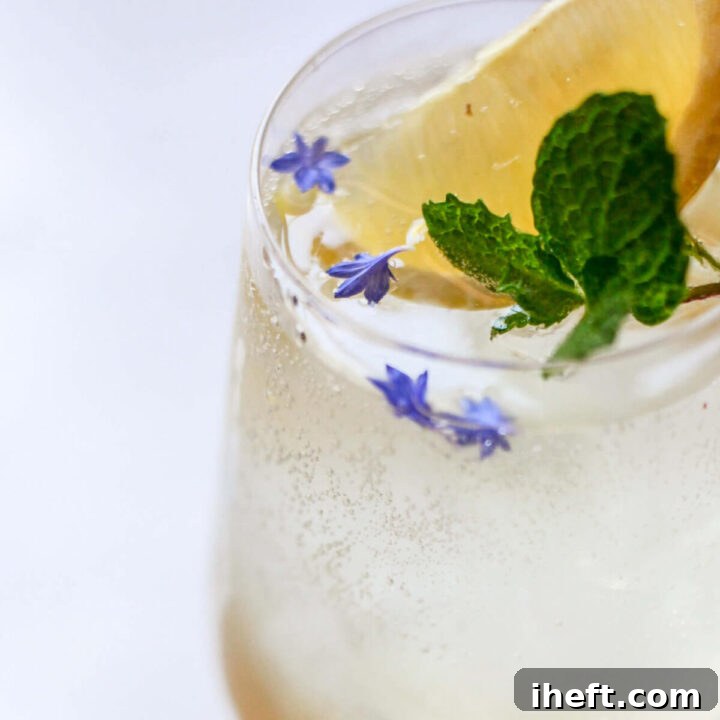 close up image of a hugo spritz in a wine glass with ice garnished with purple flowers, mint, and lemon