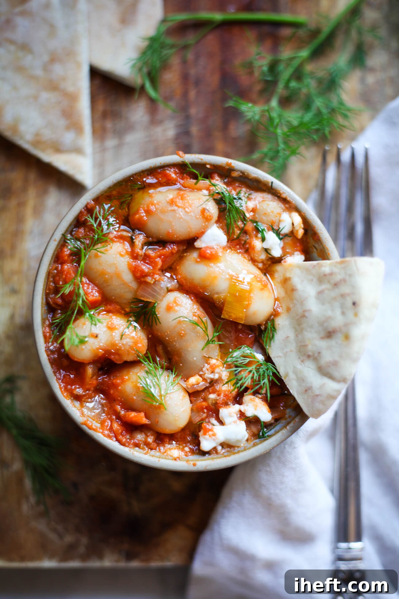 A beautifully arranged overhead photo of a rustic bowl filled with vibrant Greek giant beans in a rich red tomato sauce (Gigantes Plaki), elegantly garnished with fresh dill and crumbled feta cheese, accompanied by warm pita bread for dipping.