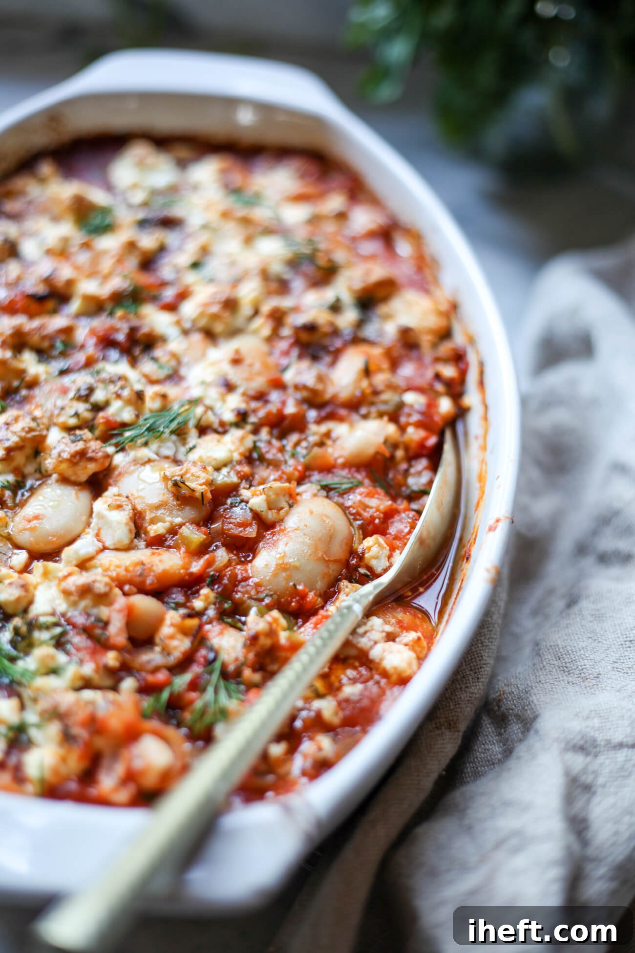 A white oval casserole dish brimming with perfectly cooked Greek baked beans (Gigantes Plaki) in a rich tomato sauce, generously topped with fresh herbs and crumbled feta cheese, ready to be served.