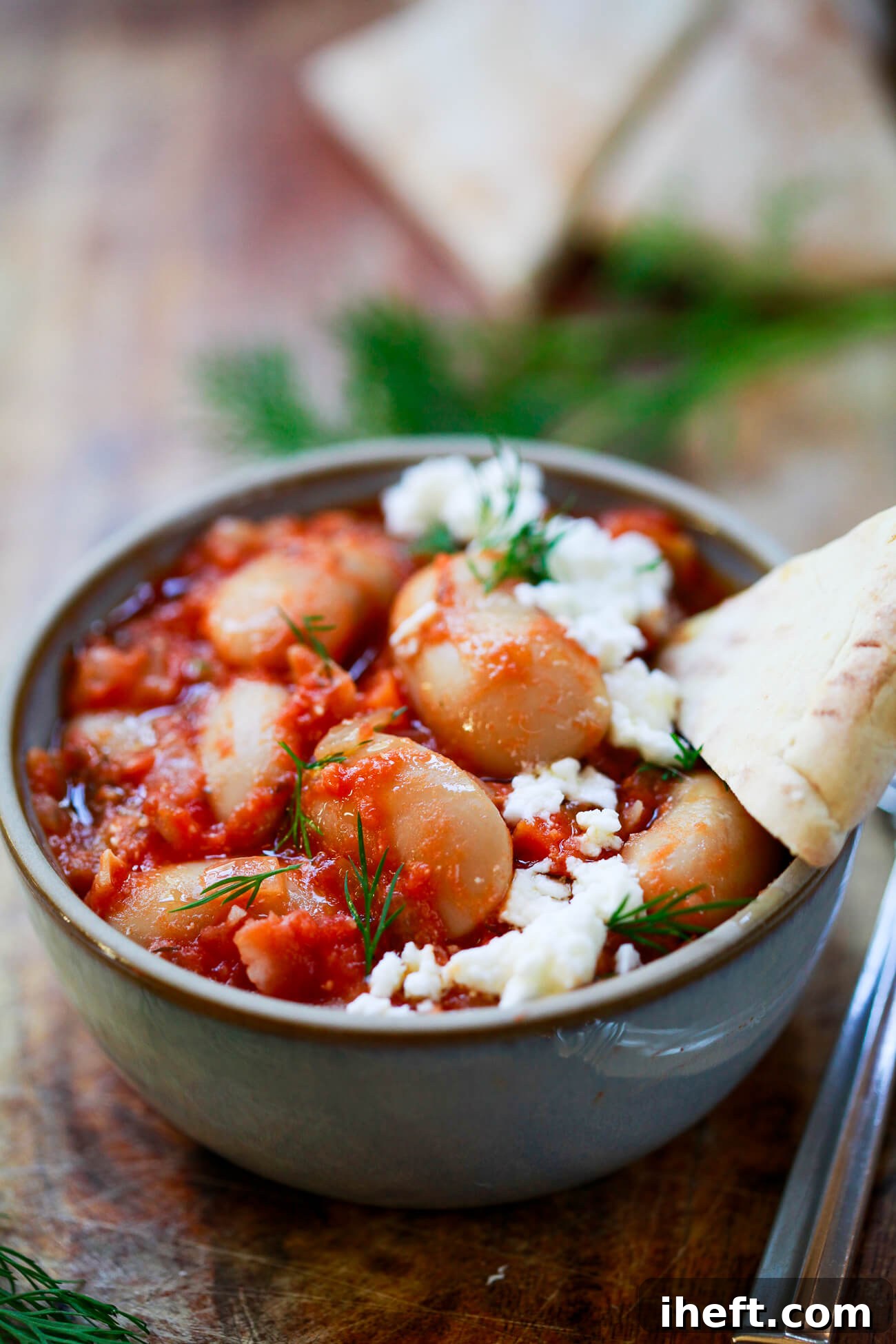A small, elegant grey bowl meticulously filled with delectable Gigantes Plaki—giant Greek white beans luxuriating in a rich tomato sauce, beautifully garnished with fresh dill and crumbled feta. A triangle of warm pita bread rests invitingly on the side.