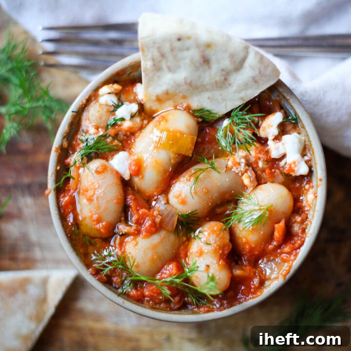 An overhead photo of a rustic bowl of Greek giant beans in a rich tomato sauce with crumbled feta and fresh dill. Warm pita bread is served on the side, inviting you to dip into the deliciousness.