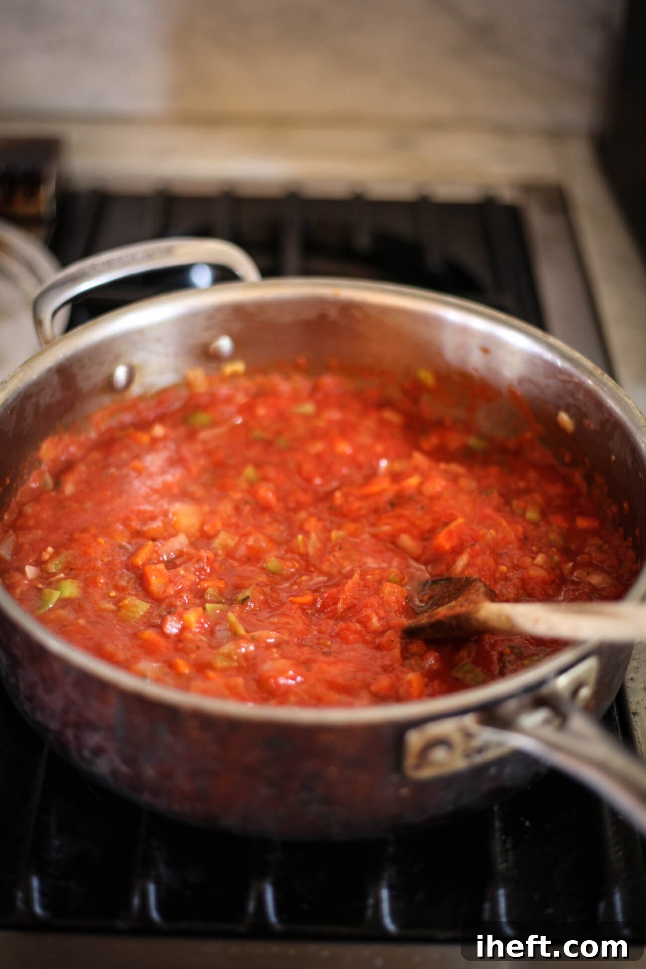 Rich tomato sauce with sautéed vegetables simmering in a large skillet on a gas stove, stirred with a wooden spoon, forming the heart of the Gigantes Plaki.
