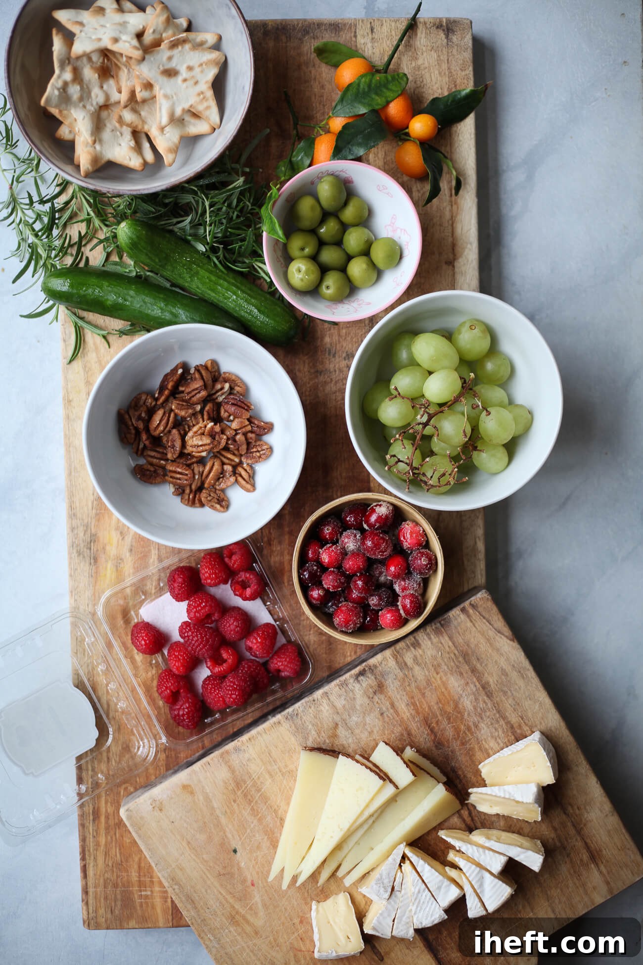 Assorted ingredients for a Christmas tree charcuterie board, including star crackers, pecans, brie, manchego, grapes, olives, cucumbers, kumquats, raspberries, cranberries, and rosemary on wooden boards, prepped for assembly.