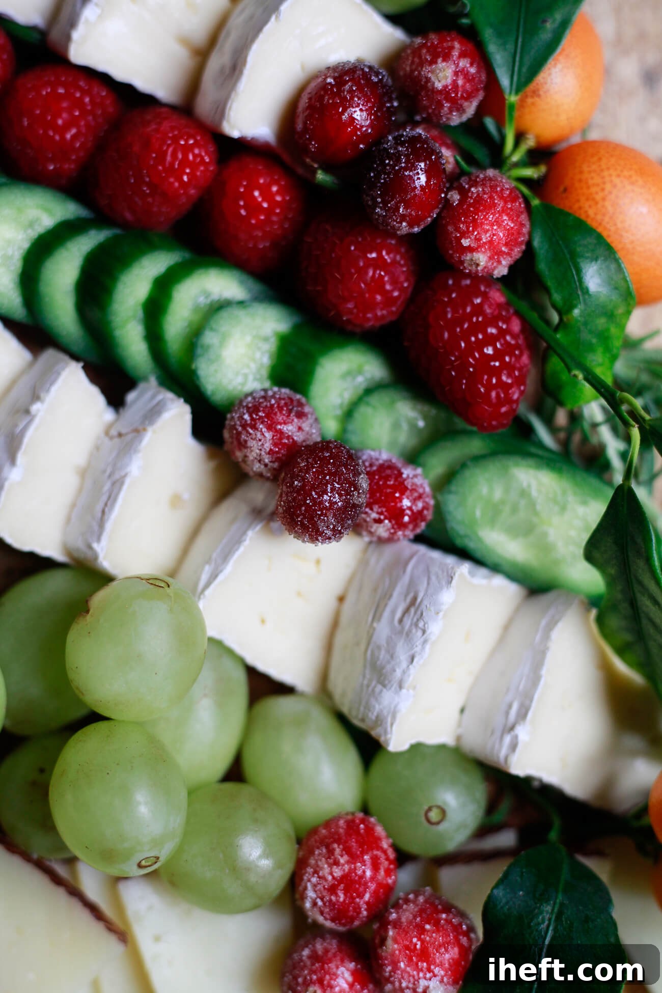 Close-up view of brie cheese slices, green grapes, cucumbers, raspberries, sugared cranberries, and kumquats arranged for a festive Christmas tree charcuterie board, highlighting fresh ingredients.