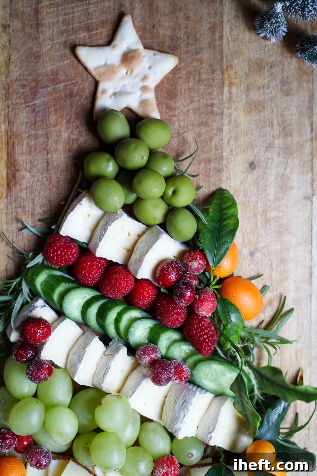 Top section of a Christmas tree charcuterie board featuring a star-shaped cracker, green grapes, brie cheese, raspberries, cucumbers, kumquats, and rosemary garnish, showcasing the festive details.