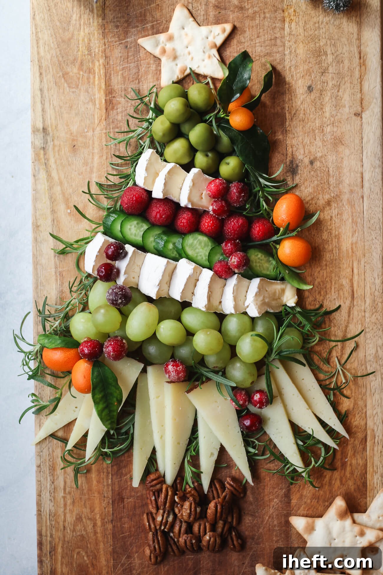 Festive charcuterie board arranged in the shape of a Christmas tree with brie, manchego, grapes, cucumbers, raspberries, kumquats, pecans, rosemary, and a star-shaped cracker topper, glistening under festive lights.