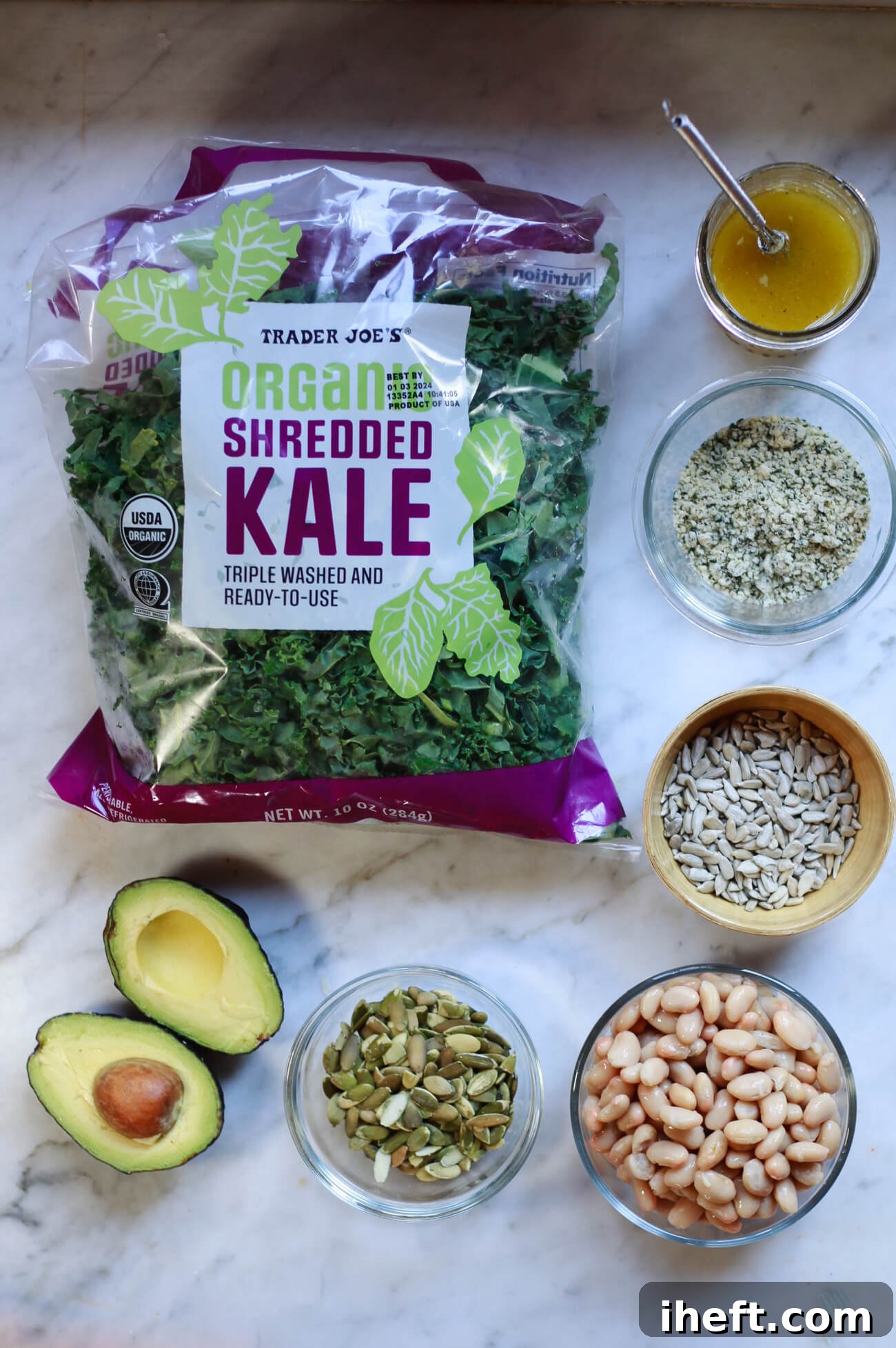 Trader Joe's shredded kale bag, homemade dressing, hemp seeds, sunflower seeds, white beans, pepitas, and avocado on a marble countertop. These are the ingredients for Erewhon Kale White bean salad. 