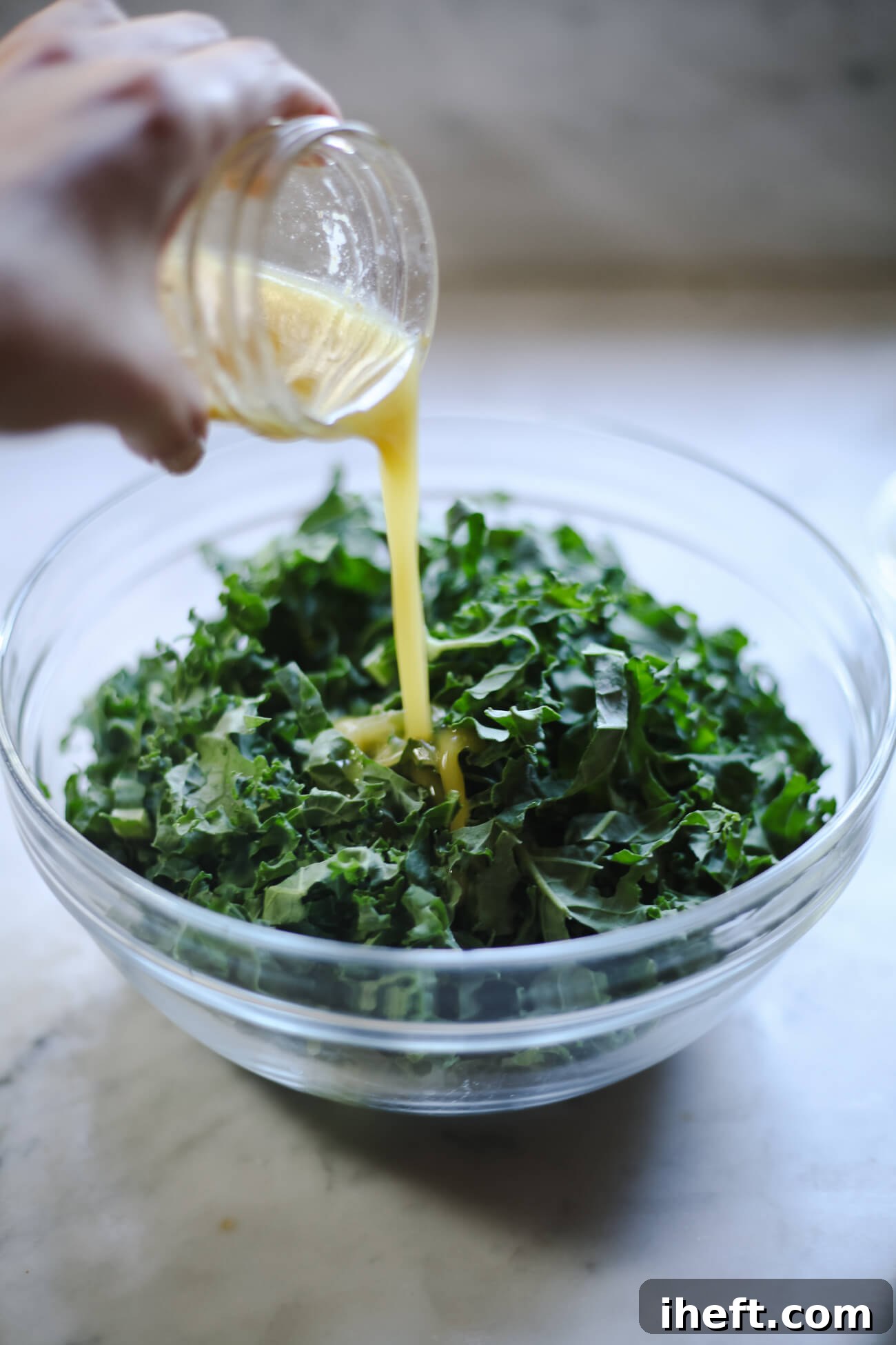 A hand pours a small jar of homemade salad dressing over shredded kale. 