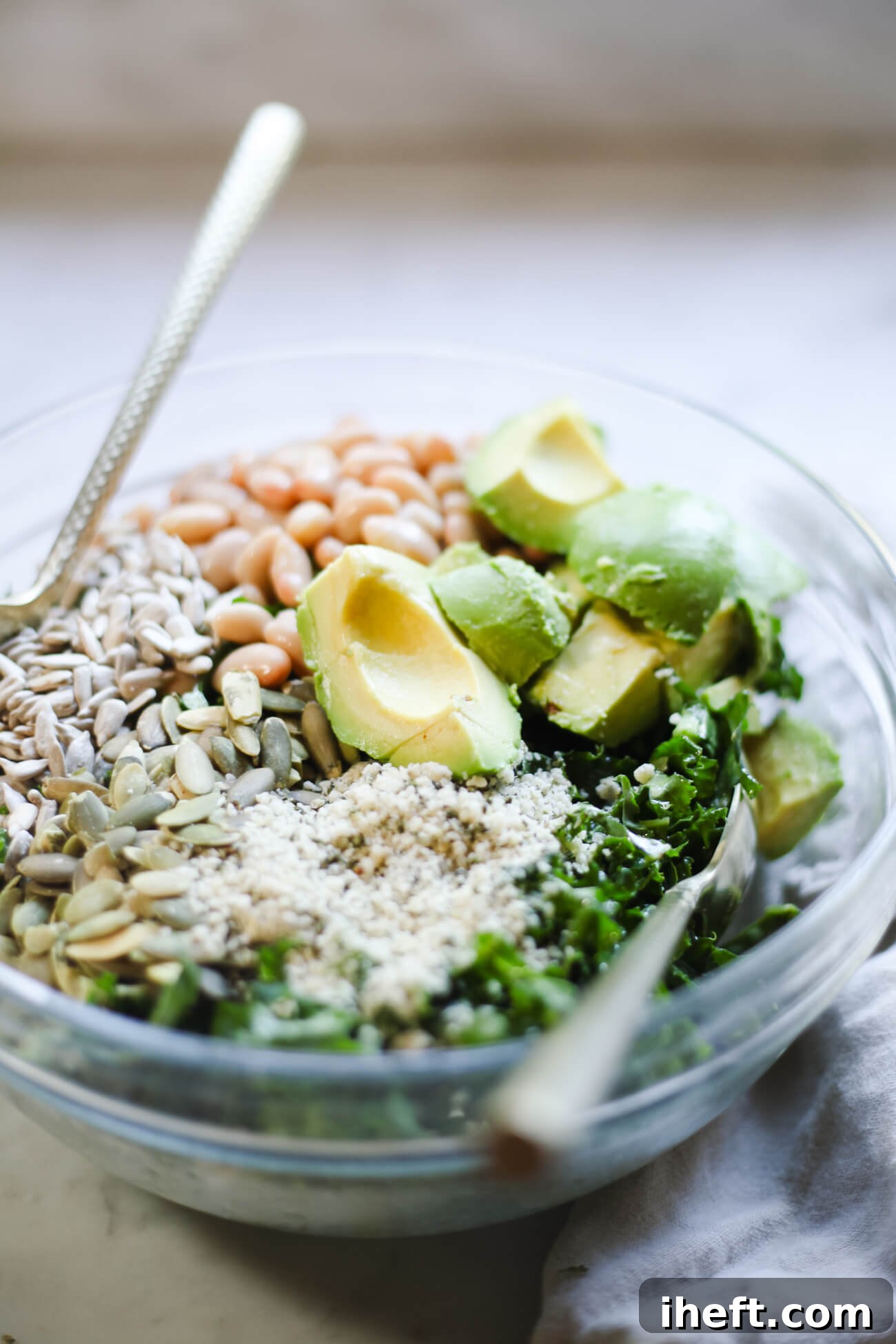 A glass bowl filled with a homemade Erewhon-inspired kale salad topped with pumpkin seeds, hemp seeds, sunflower seeds, white beans, and avocado. 