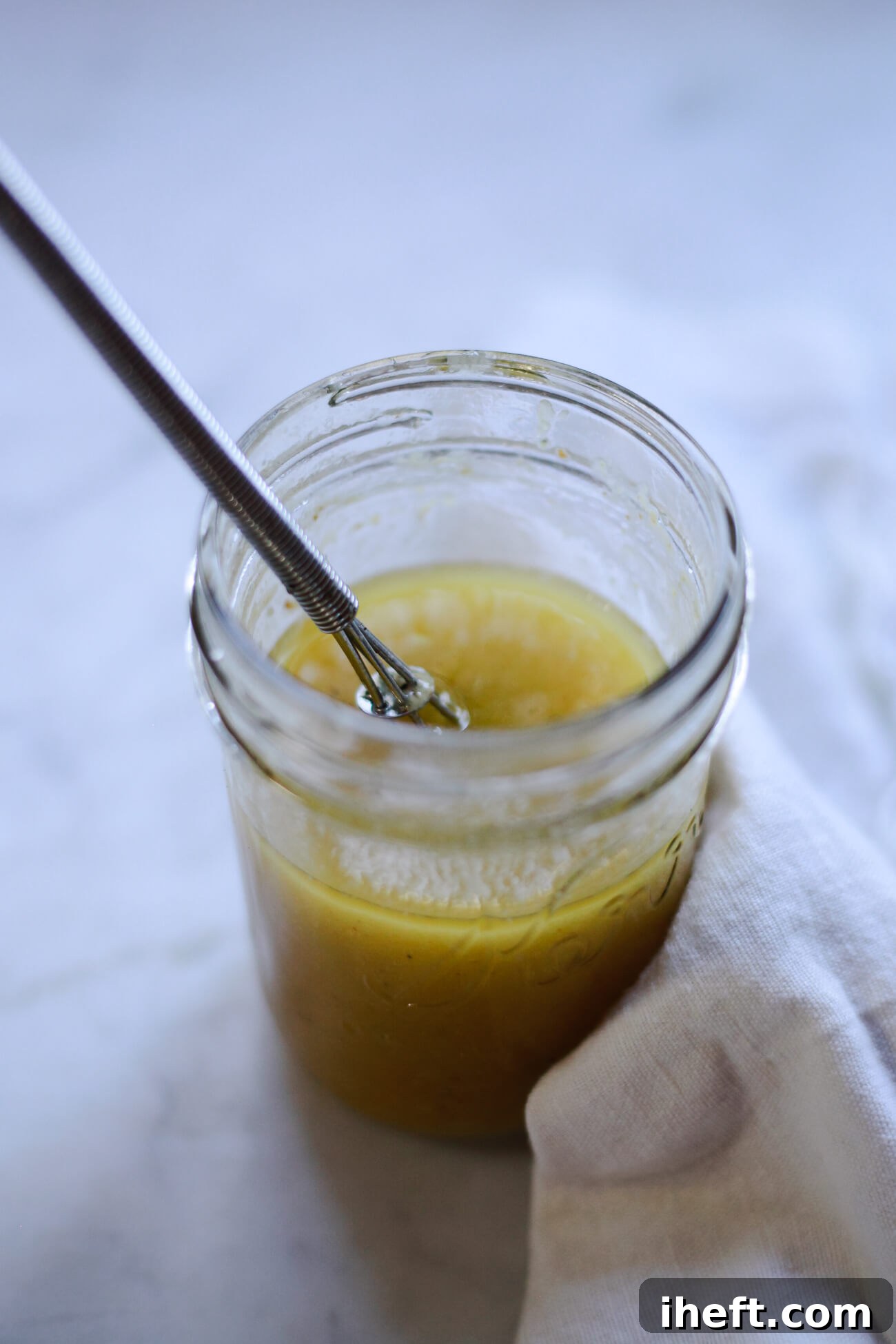 A small jar filled with a homemade lemon Dijon vinaigrette with a small whisk.