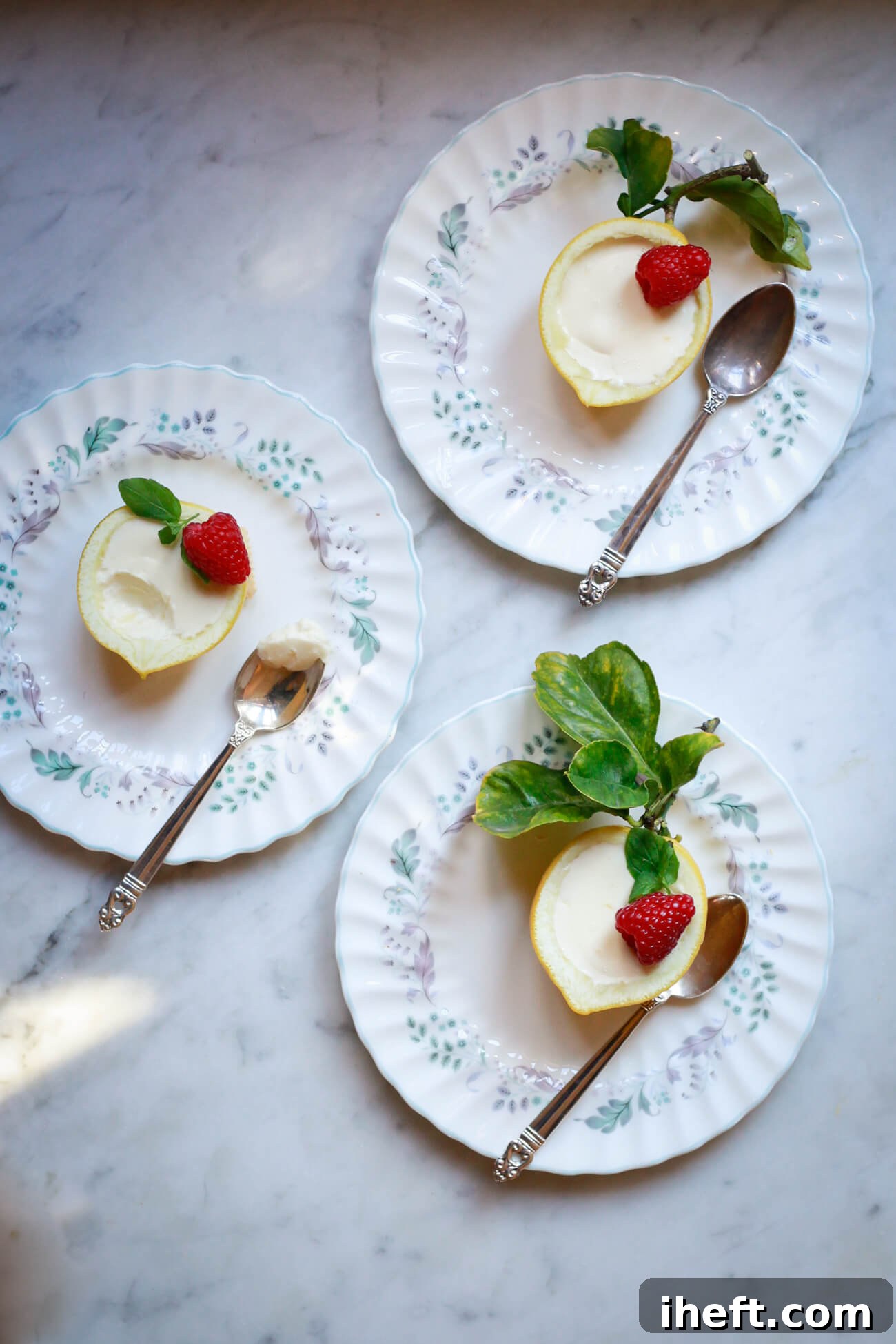 Three English china dessert plates with lemon shell halves filled with lemon posset. The cream lemon possets are garnished with raspberries and mint. The plates sit on a marble countertop. 