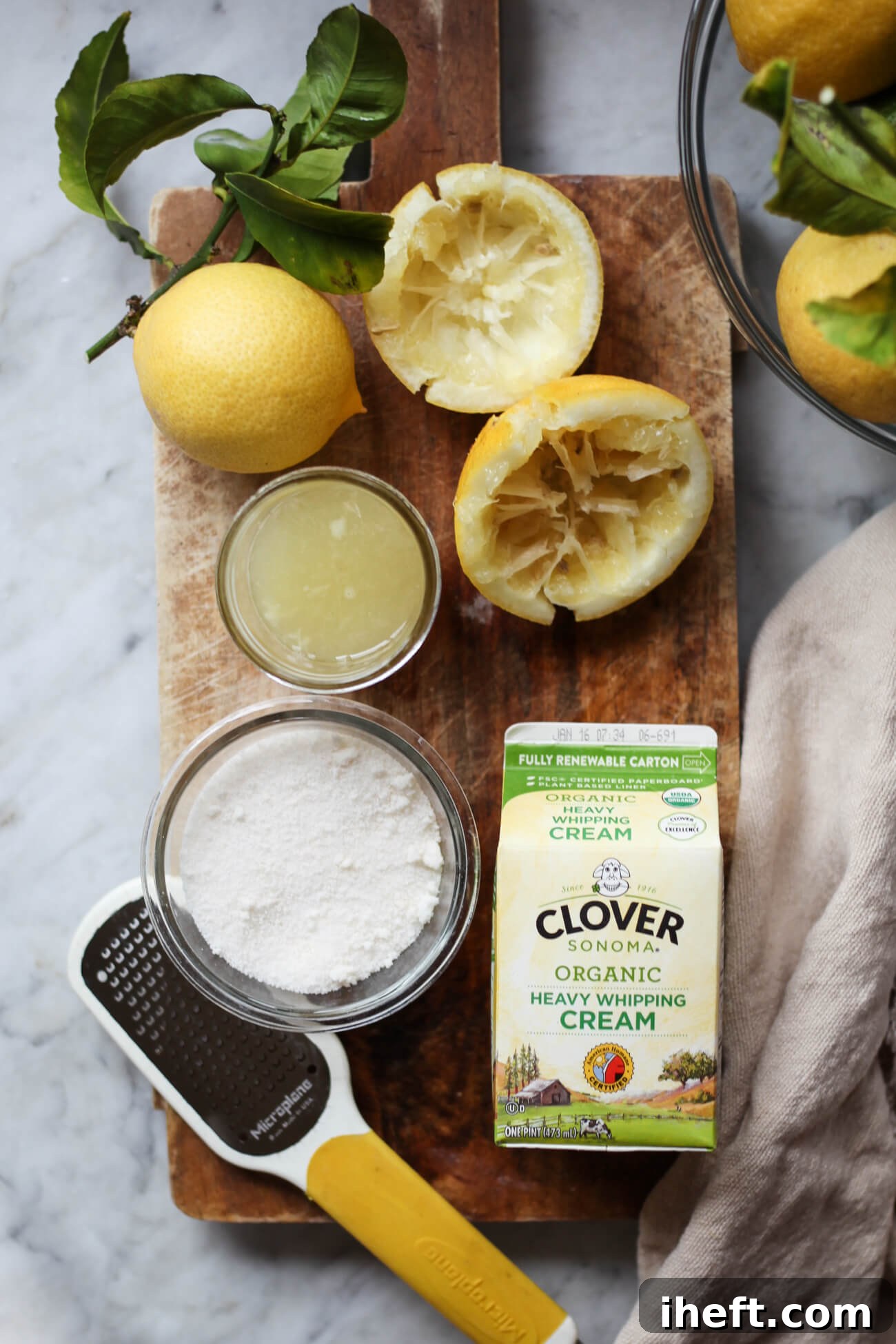Lemon posset ingredients sit on a wooden board on a marble countertop. Ingredients are lemons, fresh lemon juice, Clover organic heavy cream pint, and a bowl of sugar. 