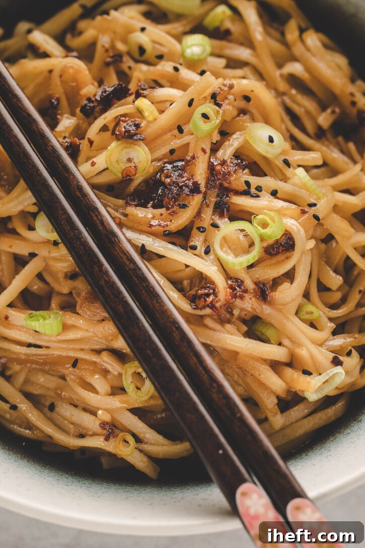 A close-up shot of a steaming bowl of homemade chili garlic noodles, generously topped with thinly sliced fresh green onions, crispy chili oil, and toasted sesame seeds. Chopsticks are poised, ready to delve into the flavorful dish, highlighting its enticing texture.