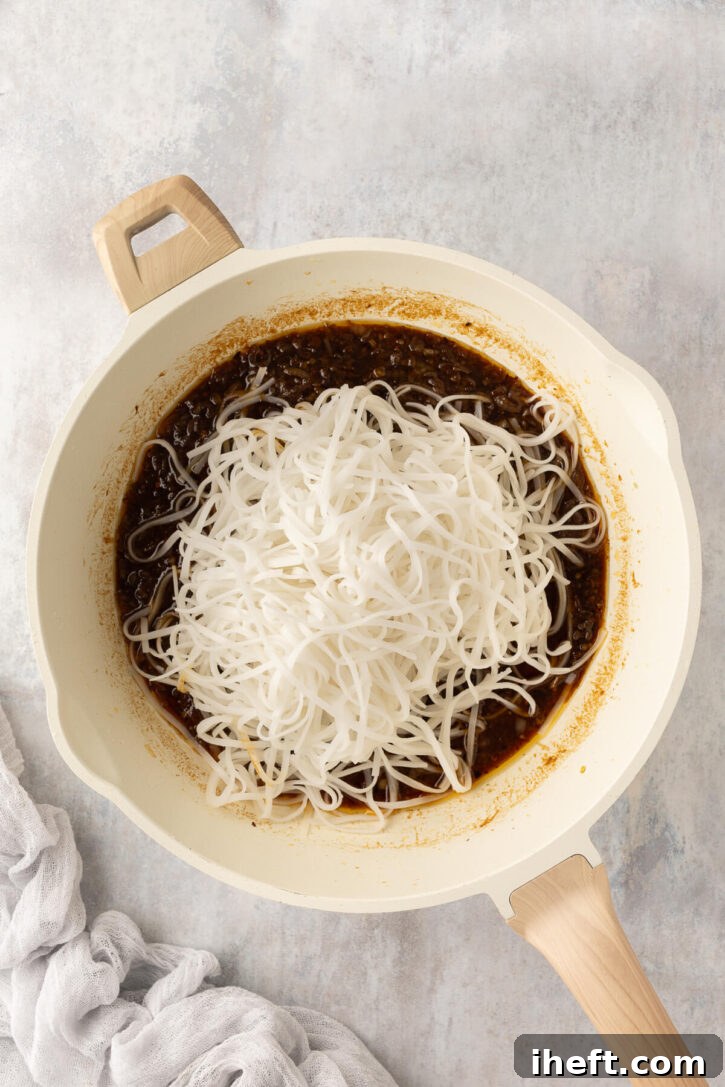 Perfectly cooked rice noodles are gently added to a large skillet, where a rich, simmering chili garlic sauce awaits, ready to be tossed together for the ultimate chili garlic noodles, ensuring every strand is coated.