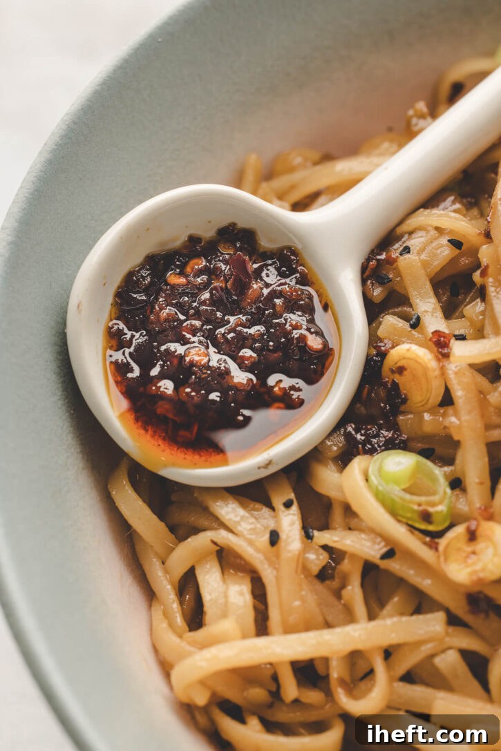 A close-up of a spoon generously filled with glossy chili garlic oil, perfectly positioned over a bowl of enticing spicy noodles, garnished with fresh scallions and toasted sesame seeds, ready to be enjoyed as a delicious topping.