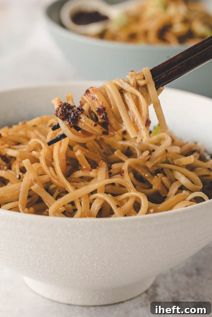 A tantalizing close-up shot of a bowl filled with chili garlic noodles, with chopsticks expertly poised to lift a perfect bite, showcasing the glossy sauce and crunchy garlic chili crisp coating each strand, inviting you to taste.