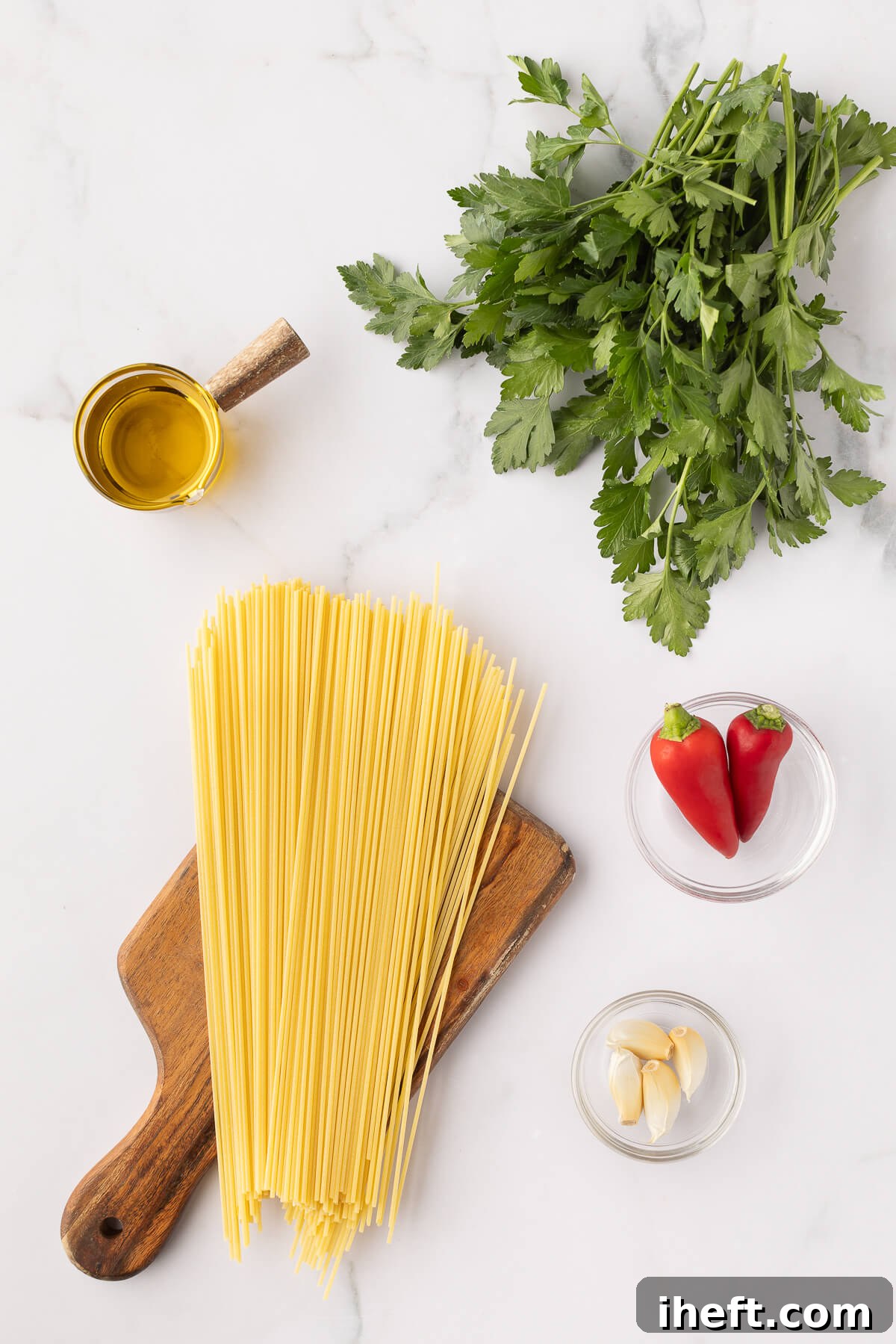 Olive oil, fresh Italian parsley, Italian peperoncino, garlic cloves, and spaghetti sit on a marble countertop. 